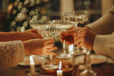 Happy friends clinking champagne glasses while celebrating New Years Eve or Christmas, sitting at festive dinner table with traditional food and burning candles